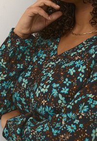 Blouse à imprimé floral avec un fond sombre, ornée de motifs turquoise, marron et crème. Manches longues froncées avec détail de boutons.