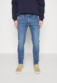Homme portant un jean slim bleu, des baskets beige et un sweat-shirt bleu marine foncé, debout devant un fond clair uni.
