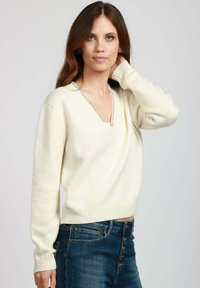 Giovane donna con lunghi capelli castani, che indossa un maglione beige a scollo a V e jeans azzurri a vita alta con bottoni, mentre posa con una mano vicino al collo.