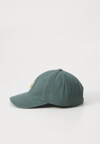 Grønn bomullsbøttecaps med buet skjerm, ventilasjonshull og et lite logomerke på siden. Enkel, strukturert design.