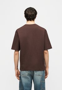 Άνδρας γυρισμένος πλάτη φορώντας απλό καφέ κοντομάνικο μπλουζάκι και δίχρωμο τζιν με πολλές τσέπες, μπροστά σε απλό φόντο.