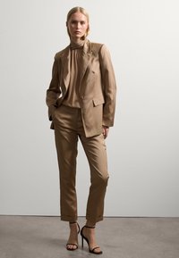 Brauner maßgeschneiderter Anzug mit einem strukturierten Blazer und einer taillierten Hose, kombiniert mit einem beigen Rollkragenpullover. Abgerundet mit schwarzen Stiletto-Absätzen.