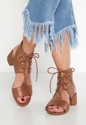 Braune Leder-Sandalen mit offenen Zehen und Schnürung sowie Blockabsätzen, getragen zu hellblauen Jeans mit ausgefranstem Saum und rot lackierten Fußnägeln.
