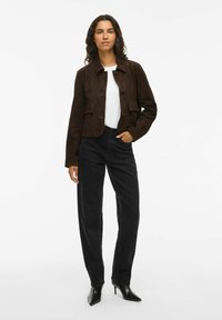 Brun ruskindjakke med et cropped design, der har to for- lommer og knaplukning, parret med højtaljede sorte jeans og sorte hæle.