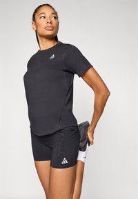 Μαύρο αθλητικό πουκάμισο με κοντά μανίκια και ραμμένες λεπτομέρειες, συνδυασμένο με μαύρο σορτς που φέρει τριγωνικό λογότυπο. Γκρι αθλητικά παπούτσια με υφή στη σόλα.