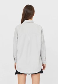 Lichtgrijs overhemd met een puntkraag en een achterplooi; heeft lange mouwen met knoopsluitingen; gedragen over een zwarte gerimpelde rok.