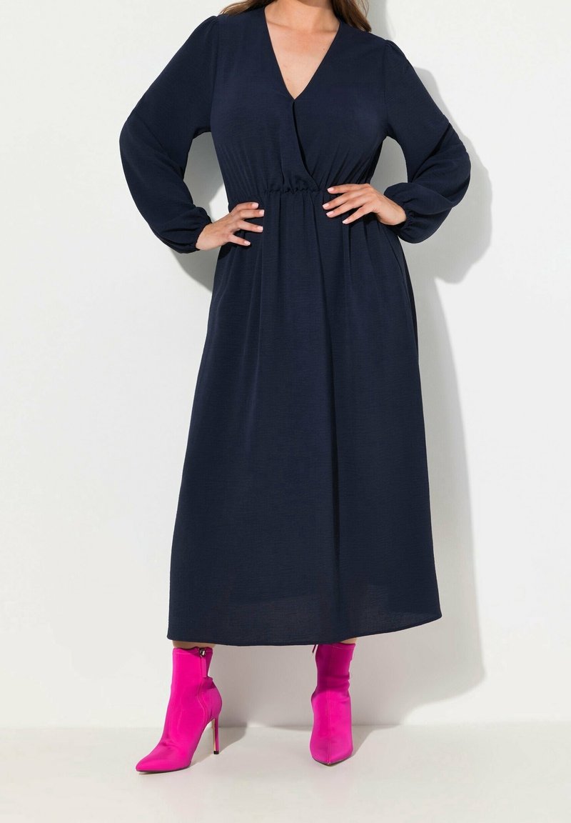 Femme portant une robe midi bleu marine à manches longues avec taille élastique et bottines à talons hauts rose vif, mains sur les hanches.