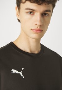 T-shirt atlético preto com decote redondo, apresentando um logo da Puma branco no lado esquerdo do peito. O tecido tem uma textura sutil.