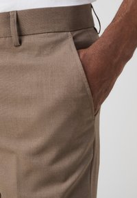 Pantaloni marroni in tessuto liscio, con tasche laterali e una vestibilità slim. La vita è piatta con una sottile cucitura decorativa.