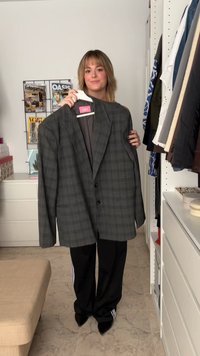 Siva karo plašč z vzorcem tartan, ki je čez velik in ima dve črni gumbi, obešen na obešalniku. Ima široke reverje in strukturirane rame.