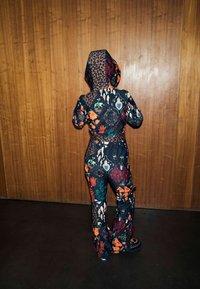 Mintás anyagból készült overall, sötét tónusokkal, élénk virág-, állatmintákkal és texturált kapucnival. Kiemelkedő részletek közé tartoznak a zsebek.