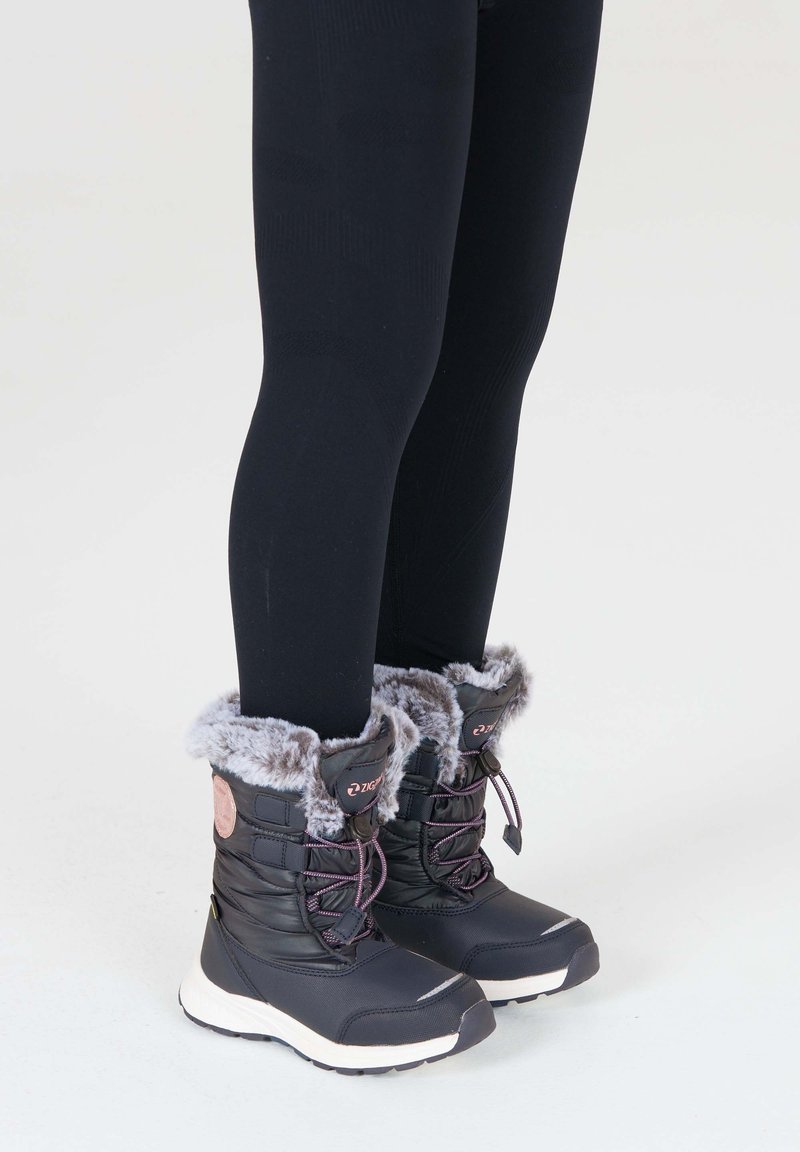 Sorte vandtætte vinterstøvler med kunstpelskant, snørebåndsdesign og kontrastfarvede hvide såler. Parret med sorte leggings.