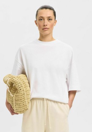 Witte oversized katoenen T-shirt met korte mouwen, gecombineerd met lichtbeige broek met trekkoord. Model houdt een gevlochten strooien tas vast.