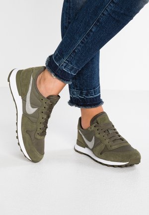 Person iført olivengrønne Nike sneakers med hvide såler og sølvfarvede swoosh-logoer, kombineret med mørkeblå optrukne jeans.