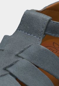 Κοντινό πλάνο σε λουρί πέδιλου από μπλε σουέντ με ορατό ράψιμο και πορτοκαλί πάτο που εμφανίζει μερικό κείμενο λογότυπου.