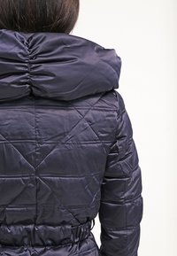 Marineblå quiltet pufferjakke med indsnøret krave, elastisk samling ved hætten og bælte i taljen. Glat tekstur og geometrisk quiltning.