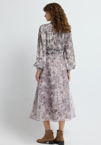 Mujer con cabello rizado que lleva un vestido midi floral de manga larga y botas marrón hasta el tobillo, de espaldas contra un fondo neutro.