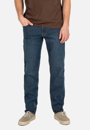 Man wearing dark blue straight-leg jeans, brown T-shirt, and beige sneakers, standing with one hand in pocket against plain background.