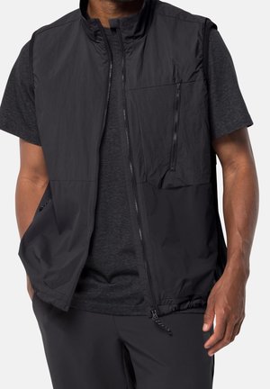Mann trägt eine schwarze ärmellose Weste mit Reißverschluss über einem dunkelgrauen T-Shirt mit kurzen Ärmeln, die Hände teils in den Westentaschen.