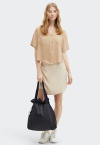 Czarna torba tote z nylonu z zebranym wierzchem, gładką teksturą i detalem logo. Modelka nosząca jasnobeżową koszulę z krótkim rękawem i spódnicę.