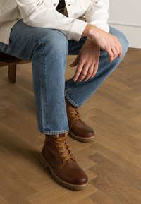 Braune Leder-Stiefeletten mit runden Zehen und Schnürsenkeln, kombiniert mit hellblauen Denim-Jeans, auf einem Holzboden sitzend.