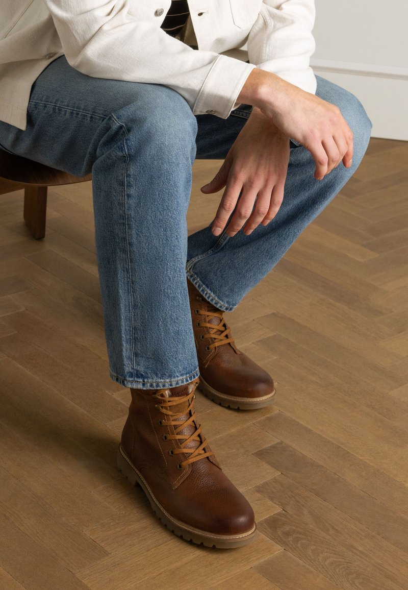 Braune Leder-Stiefeletten mit runden Zehen und Schnürsenkeln, kombiniert mit hellblauen Denim-Jeans, auf einem Holzboden sitzend.
