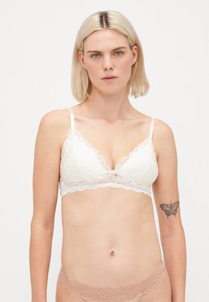 Blonde vrouw met blauwe ogen, gekleed in een witte kanten bralette en beige kanten ondergoed, staande tegen een eenvoudige witte achtergrond.
