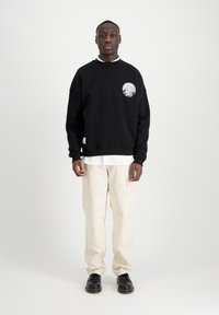 Sweatshirt nero oversize con design grafico sul lato sinistro del petto, abbinato a pantaloni cargo beige e scarpe nere, su uno sfondo neutro.