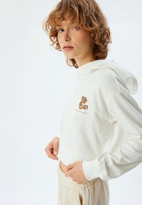Weißes, kurzes Hoodie aus weichem Stoff, mit einer braunen Teddybär-Stickerei auf der Brust. Dazu passende Sweatpants in einem hellen Beige.