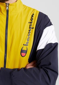 Cortavientos con bloques de color en amarillo, azul marino y blanco. Presenta un cuello alto, cierre completo y un prominente logo negro en la parte frontal.