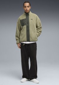 Giacca in fleece verde oliva con una tasca anteriore verde scuro. Presenta un colletto alto, chiusura con zip e una superficie testurizzata. Abbinata a pantaloni neri.