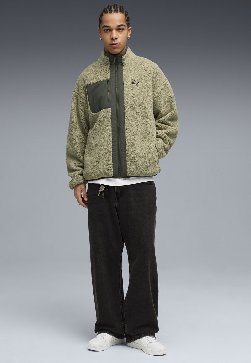 Giacca in fleece verde oliva con una tasca anteriore verde scuro. Presenta un colletto alto, chiusura con zip e una superficie testurizzata. Abbinata a pantaloni neri.