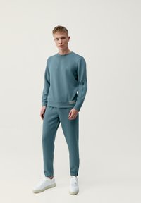 Svetlo modrá mikina a zodpovedajúce nohavice vyrobené z mäkkej látky. Zipsy na rukávoch a textúrovaná nášivka na páse. Biele topánky.