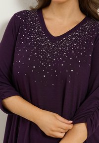 Lila V-ringad topp med trekvartsärmar har ett glittrande mönster av rhinstenar vid halsringningen. Mjuk, lätt tyg.