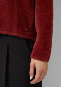 Maglione rosso morbido con una texture soffice, che presenta un orlo arrotondato e un accento con logo dorato. Abbinato a un indumento nero, evidenziando il contrasto.