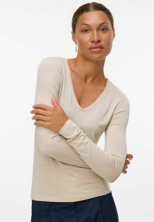 Beige langemouw V-hals top met een geribbelde textuur. Heeft aansluitende mouwen en een gladde afwerking. Gdrag met marineblauwe broek.