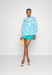 Blusa floral em azul claro com colarinho preto, combinada com calções turquesa. O modelo segura uma mala amarela texturizada e usa sandálias brancas.