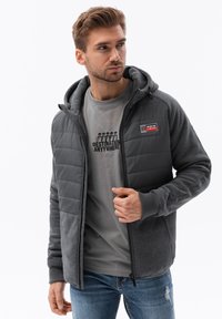 Chaqueta con capucha gris y delantero acolchado, con puños de canalé y un parche de logo. Lleva puesta una camiseta gris con un diseño impreso. Jeans de denim casual.