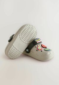 Hellgraue Clogs mit einer strukturierten Sohle, schwarzem Riemen und bunten, abnehmbaren Dekorationspins in verschiedenen Formen und Symbolen.