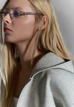 Jonge vrouw met lang blond haar, een bril met decoratieve montuur en een lichtgrijze hoodie, kijkend naar links tegen een neutrale achtergrond.
