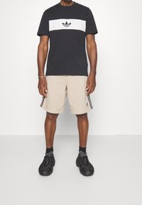 Muž oblečený v černo-bílém tričku Adidas, béžových šortkách Adidas s černými pruhy, černých ponožkách a černých teniskách stojící na bílém pozadí.