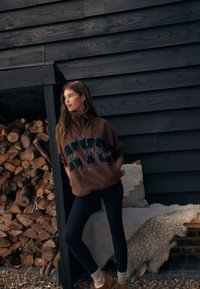 Brun fleece-pullover med høj krave og trykt "NYC" tekst, kombineret med sorte leggings og brune tøfler. Tekstureret baggrund af træstammer.