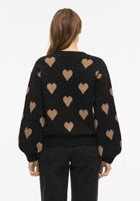 Czarny sweter z beżowym wzorem serca; posiada luźne rękawy i prążkowany pas biodrowy. Miękka tekstura, okrągły dekolt i casualowy design.