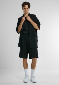 Svart ribbad kortärmad skjorta, lager-på-lager över en svart t-shirt och tillsammans med matchande shorts. Vita sneakers och strumpor fulländar looken.