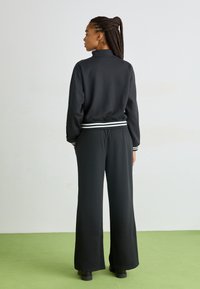 Schwarzer, lässig geschnittener Jumpsuit mit hohem Kragen, gestreiftem elastischen Taillenbund und weitem Bein. Glattes Material mit matter Oberfläche.