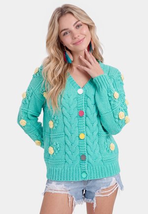 Ung kvinde med langt blondt hår iført en turkis strikket cardigan med farverige knapper og gule blomster detaljer samt denim shorts.
