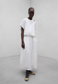 Femme grande à la peau foncée avec des cheveux tressés portant un haut blanc ample à manches courtes et une longue jupe, des ballerines beige et des chaussettes foncées, debout sur un sol gris.