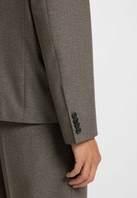 Brown textured blazer sleeve with a row of four black buttons. Tailored fit, smooth material, and clean lines visible in the design.