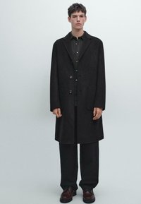 Ung mand står oprejst mod en ensfarvet baggrund iført en lang mørk frakke, matchende bukser og brune lædersko.