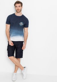 T-shirt blu navy sfumato con grafica bianca e acquamarina sul petto, abbinato a pantaloni cargo in denim scuri e sneakers bianche.